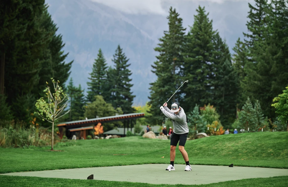 A person is swinging a golf club on a lush golf course, surrounded by trees and mountains in the background.