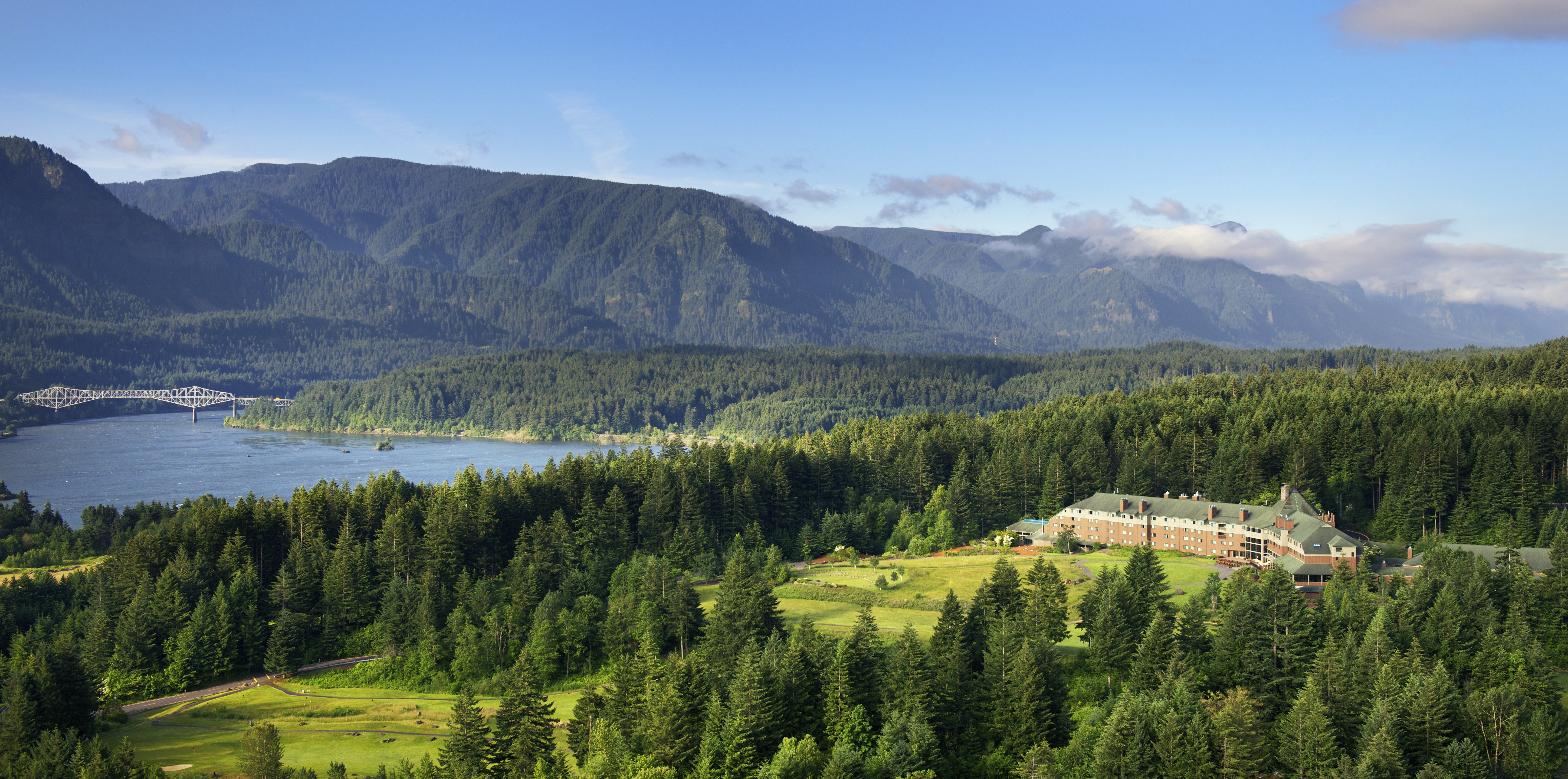 A scenic view of lush green forests, a large building, a lake, and mountains in the background under a clear blue sky.