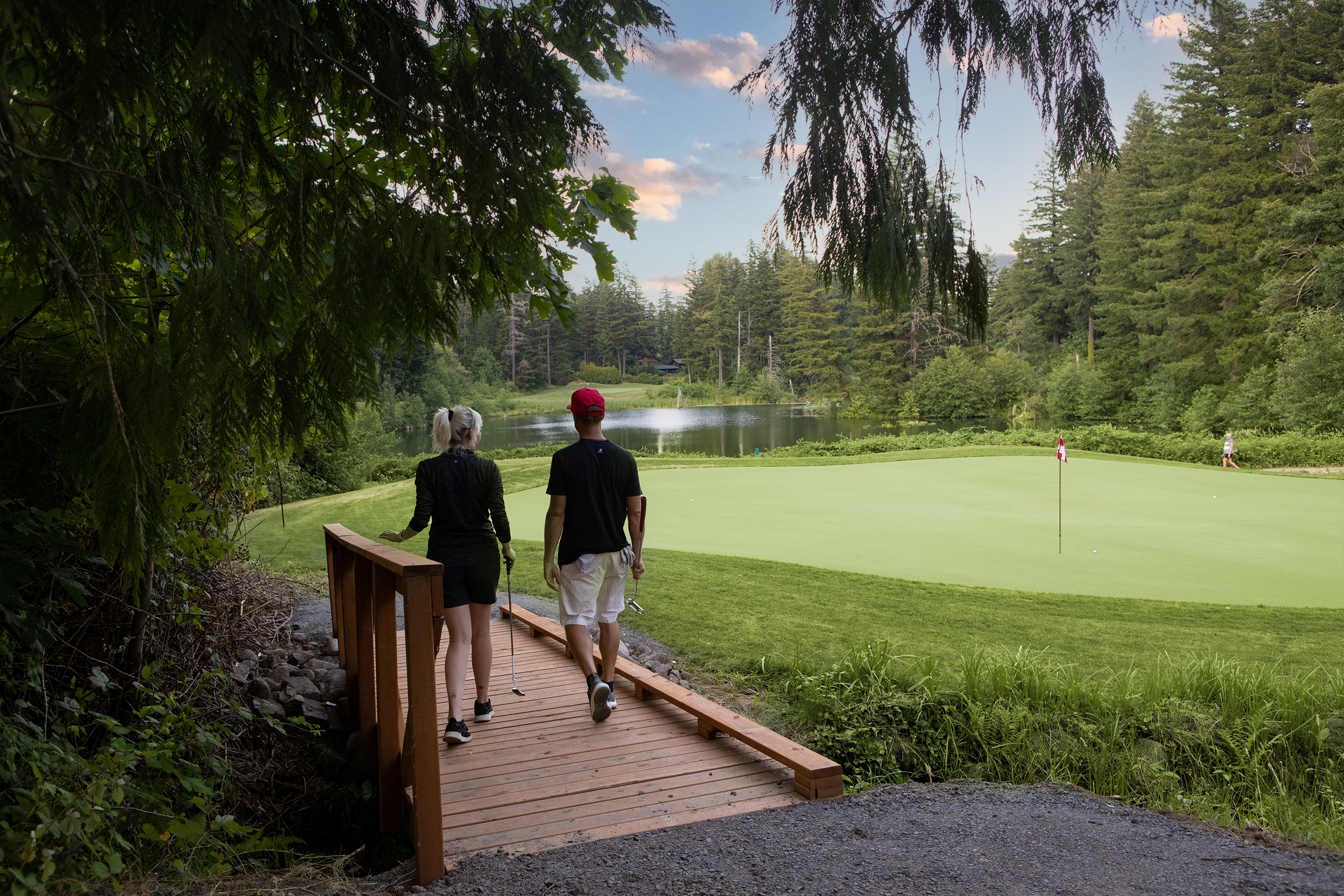 Two people walk across a small bridge towards a golf course green, surrounded by trees and a pond, under a partly cloudy sky.