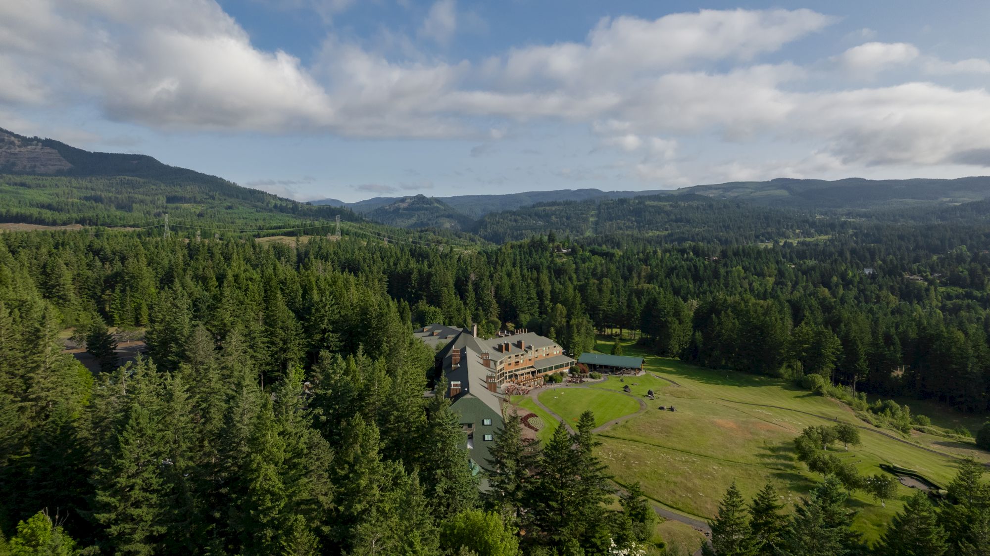 The image shows a lush, forested landscape with a building nestled among the trees, set against a background of hills and a partly cloudy sky.