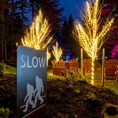 At night, a “SLOW” sign stands beside glowing light-wrapped trees and rows of chairs in a garden event area.