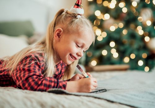 A child with a festive hat is joyfully drawing on a bed, with a blurred Christmas tree and lights in the background, creating a cozy ambiance.