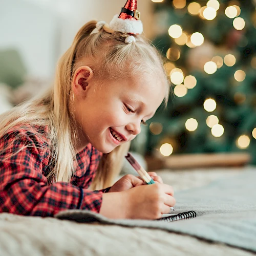 A child with a festive hat is joyfully drawing on a bed, with a blurred Christmas tree and lights in the background, creating a cozy ambiance.
