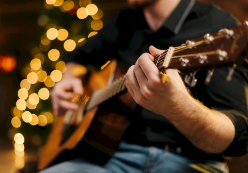 A person is playing an acoustic guitar, with blurred lights in the background creating a warm, festive ambiance.