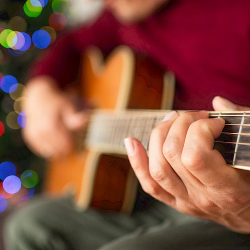 A person playing an acoustic guitar with a colorful, blurred background, possibly holiday lights, creating a warm, festive atmosphere.