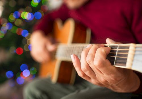 A person is playing an acoustic guitar with festive, colorful lights blurred in the background, creating a warm and cozy atmosphere.