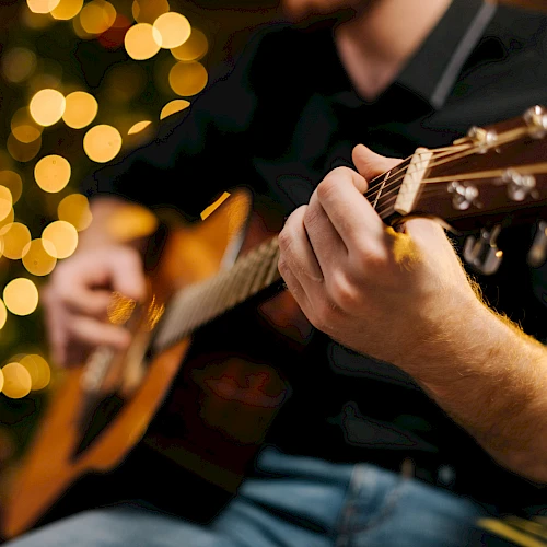 A person is playing an acoustic guitar, with blurred lights in the background creating a warm, festive ambiance.