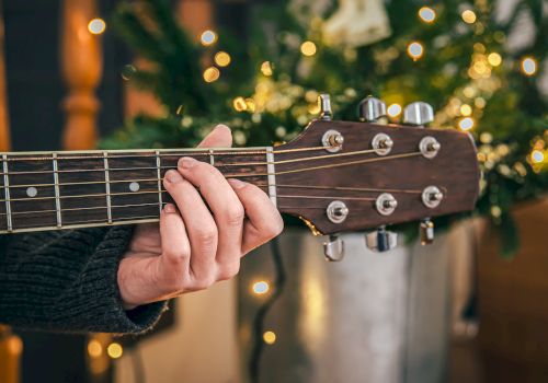 A person is playing an acoustic guitar with festive lights and a decorated tree in the background.