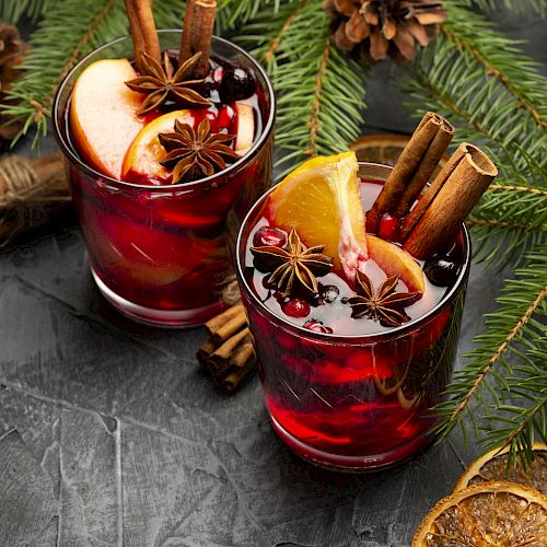 Two glasses of mulled wine with spices, fruits, and cinnamon sticks, surrounded by pine branches and cones on a dark surface.