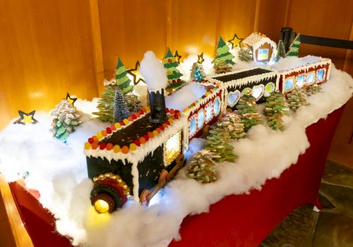 A festive gingerbread train with candy decorations, surrounded by snow and trees, sits on a table, glowing with string lights.
