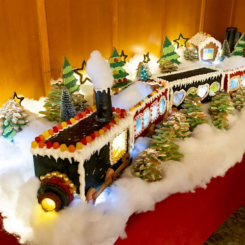 A festive gingerbread train with candy decorations, surrounded by snow and trees, sits on a table, glowing with string lights.