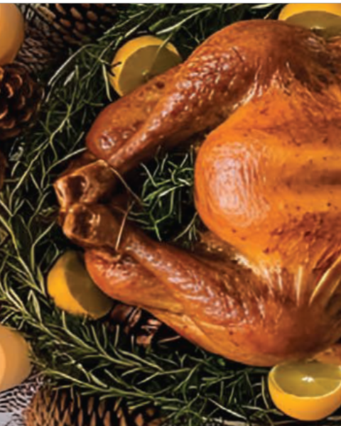A roast turkey garnished with herbs and citrus is surrounded by candles, pine cones, and a plate of roasted potatoes.