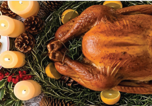 A roast turkey garnished with herbs and citrus is surrounded by candles, pine cones, and a plate of roasted potatoes.