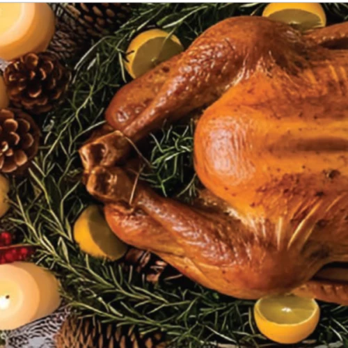 A roast turkey garnished with herbs and citrus is surrounded by candles, pine cones, and a plate of roasted potatoes.