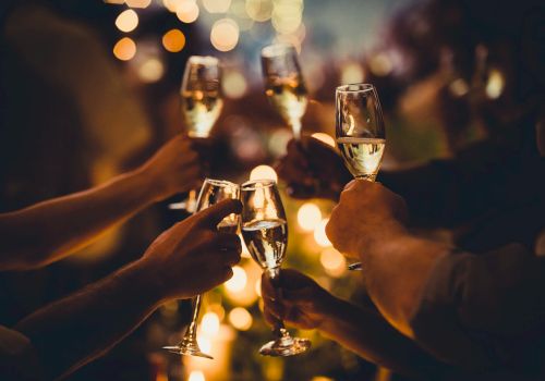 People are raising champagne glasses in a celebratory toast, with warm lighting and blurred bokeh in the background.