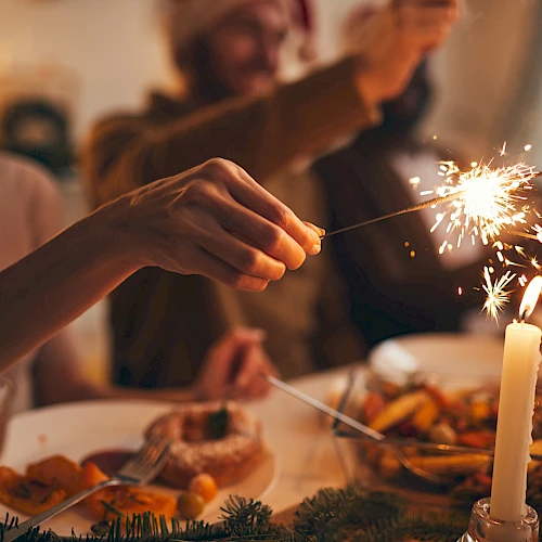 A festive dining scene with people holding sparklers at a table set with food and a lit candle.