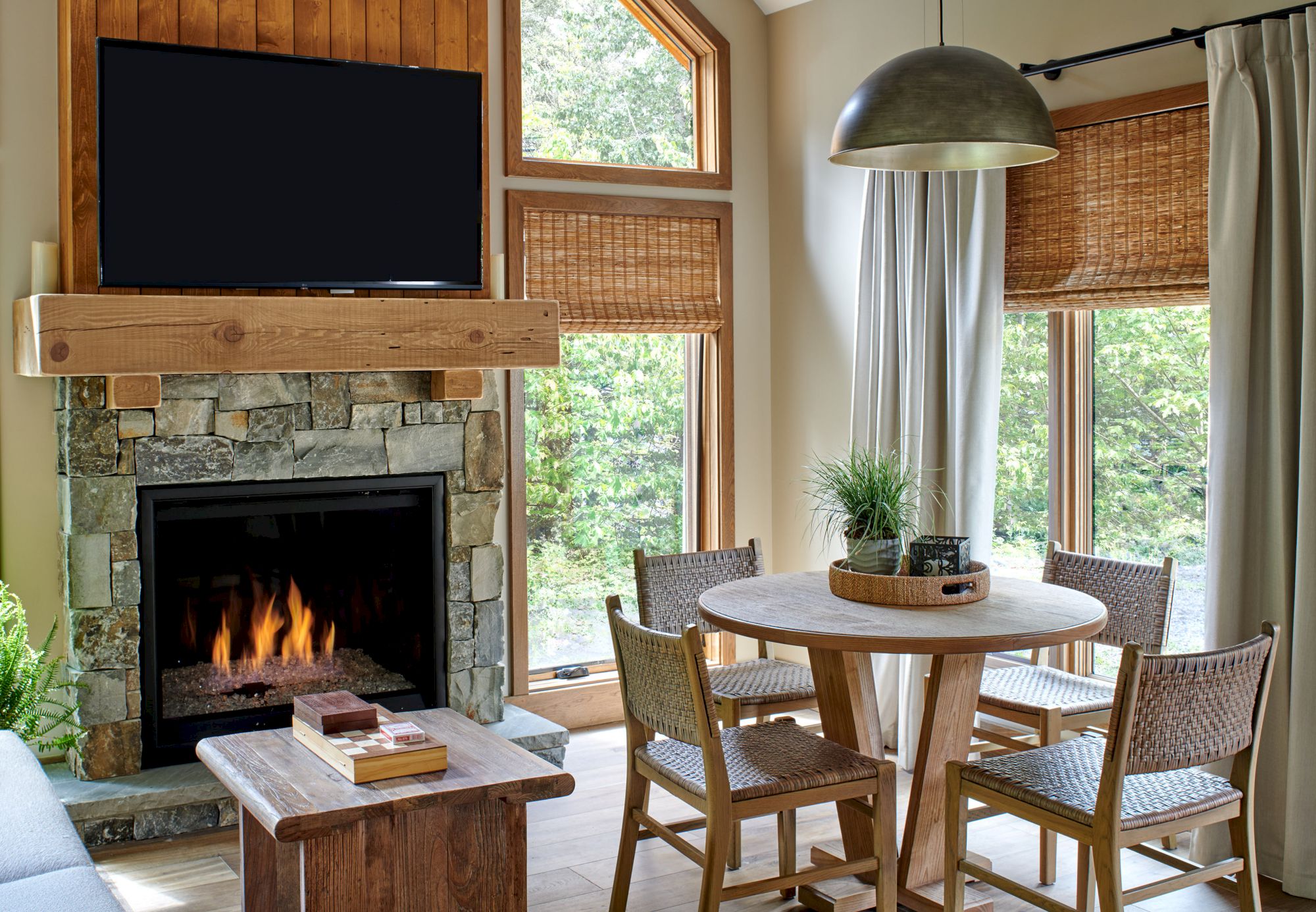 Cozy living area with a stone fireplace, wall-mounted TV, wooden table with chairs, and large window views.