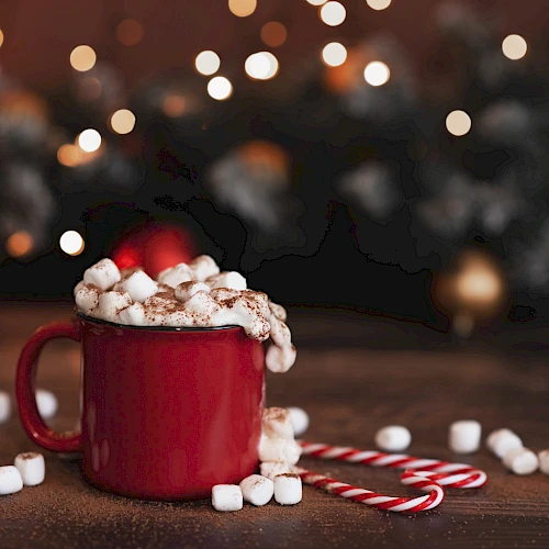 A red mug filled with hot chocolate and marshmallows, surrounded by candy canes, with a festive, blurred light background.