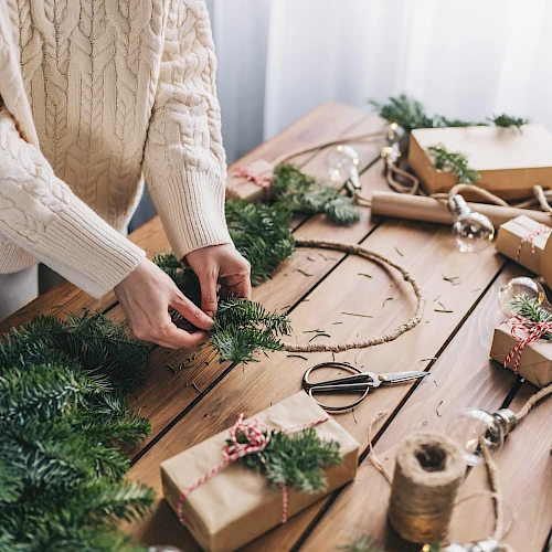 A person is crafting a holiday wreath at a table filled with wrapped gifts and greenery, creating a festive atmosphere.
