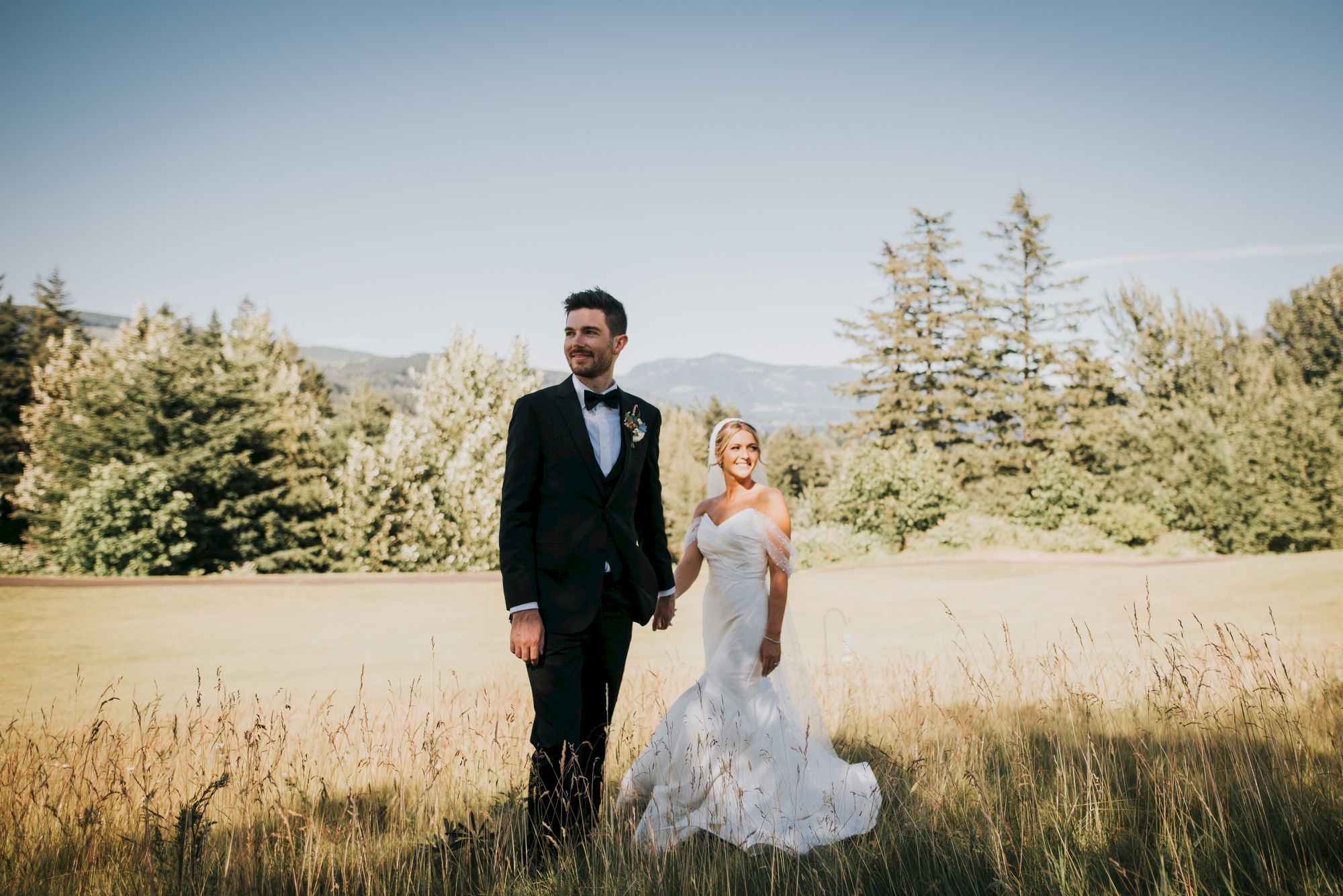 A couple in wedding attire stands in a field with trees and mountains in the background, under a clear blue sky.