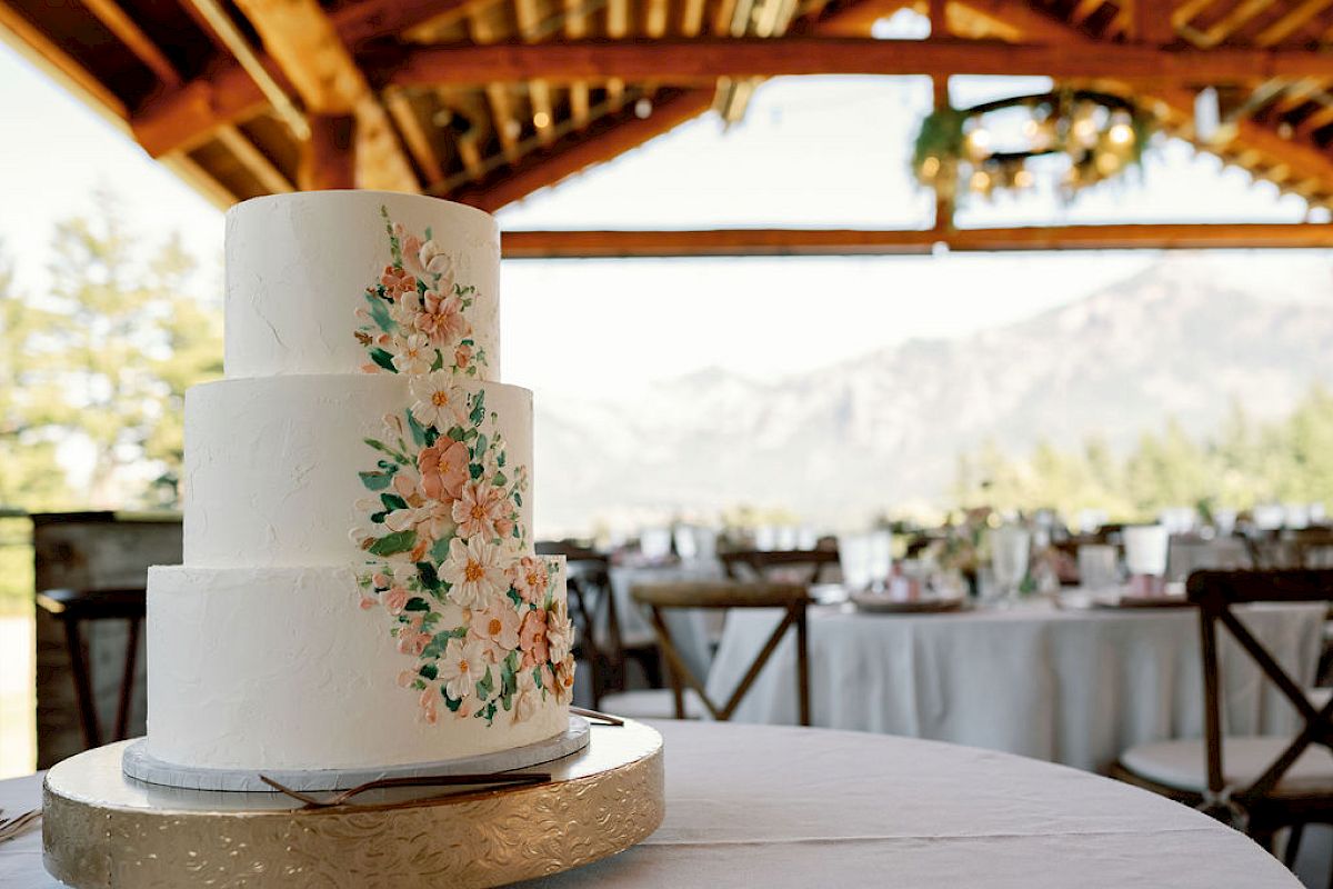 A three-tier wedding cake with floral design is set on a table under a rustic wooden pavilion, with mountains visible in the background.