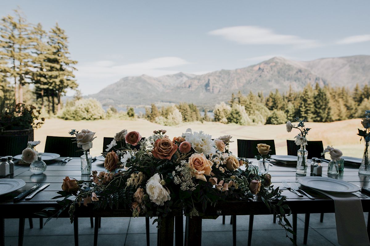 A beautifully set outdoor table with floral arrangements, overlooking mountains and a scenic landscape under a clear sky.