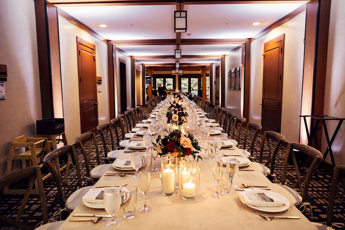 A long, elegantly set dining table with candles and floral centerpieces inside a well-lit hall, ready for a formal event or gathering.