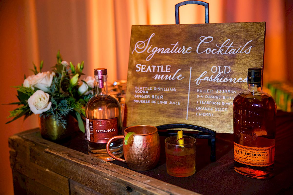 A cocktail menu displays "Seattle Mule" and "Old Fashioned" beside bottles and flowers on a wooden surface.