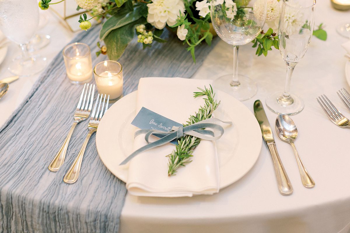 A beautifully set table with a white plate, napkin, rosemary sprig, cutlery, candles, and a floral centerpiece.