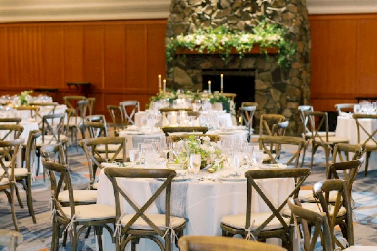 A decorated event space features round tables with white tablecloths, elegant centerpieces, and wooden chairs near a stone fireplace.