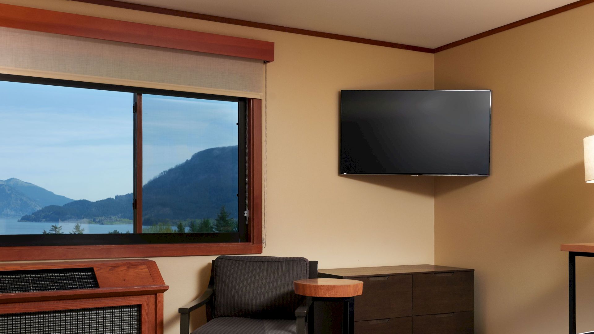 A cozy hotel room features a bed, TV, desk, lamps, and a large window with a serene view of mountains and water outside.