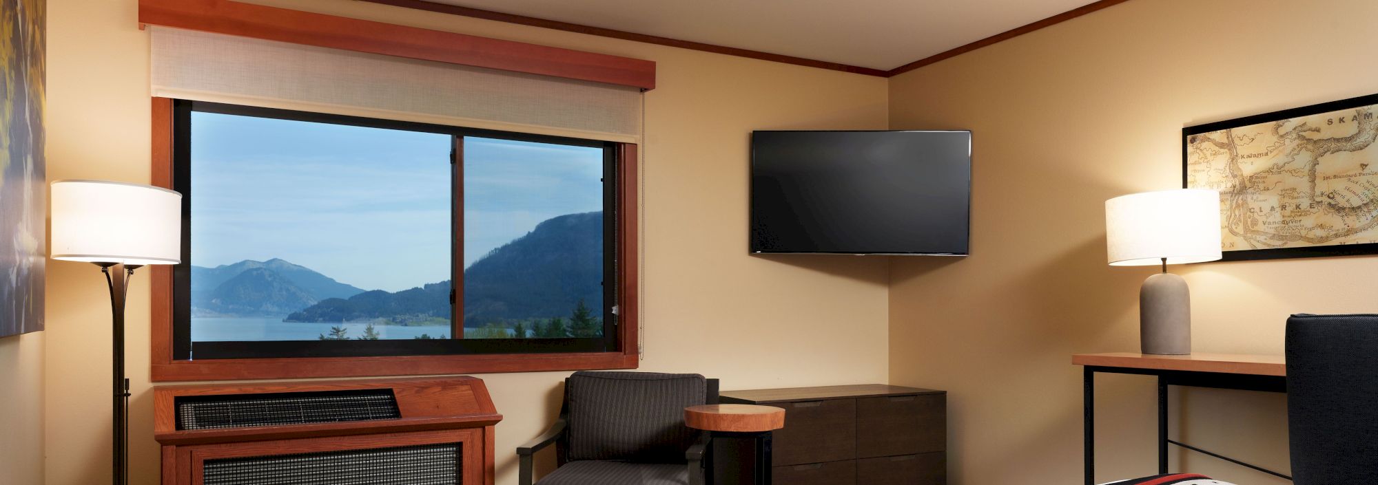A cozy hotel room features a bed, TV, desk, lamps, and a large window with a serene view of mountains and water outside.