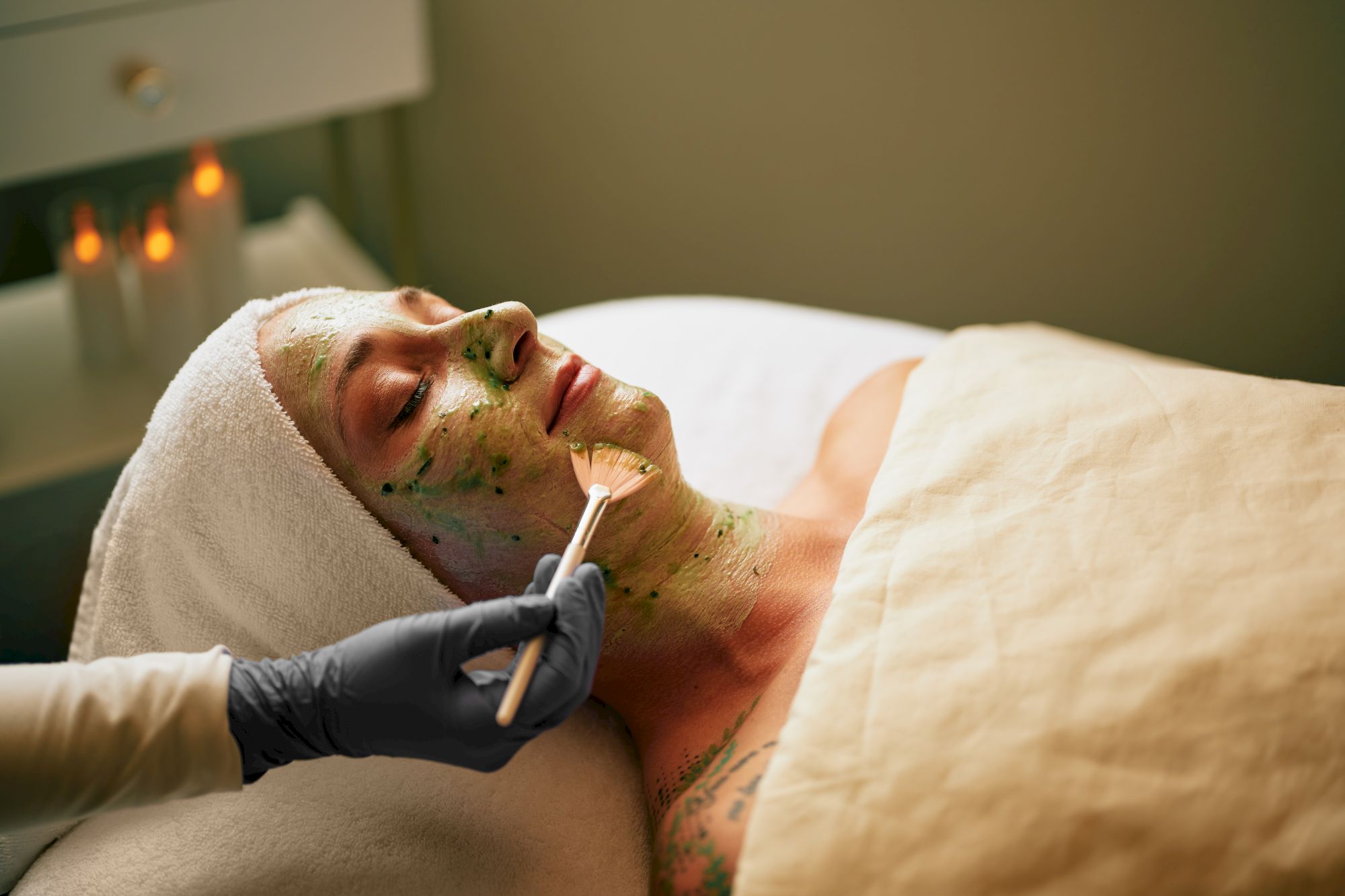 A person receiving a facial treatment with a green mask being applied by an attendant wearing gloves, while lying under a blanket.