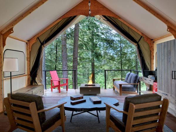 A cozy cabin living area with a round coffee table, four wooden chairs, a fire pit in a forested tented cabin, and red and gray chairs on a deck outside.