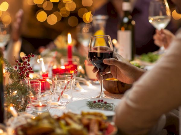 A festive dinner scene with wine glasses, candles, and soft bokeh lights, as someone toasts with a glass of red wine.