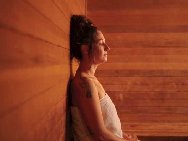 A person is sitting in a wooden sauna, wrapped in a towel, with eyes closed and head resting on the wall.