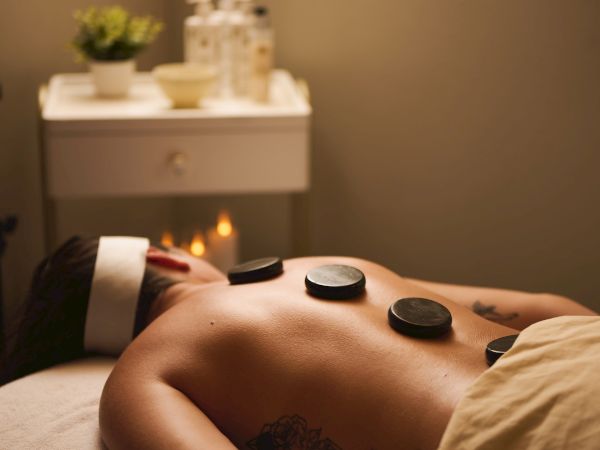 A person receiving a hot stone massage on a massage bed, with a towel and soothing room setup, featuring a small table with supplies nearby.