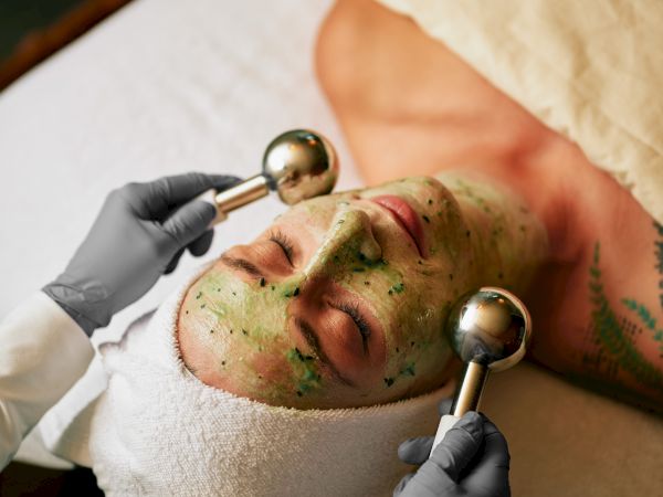 A person receiving a facial treatment with a green face mask and metal rollers, wearing a towel on their head, in a spa setting.