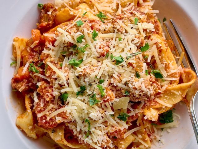 A plate of pasta—tagliatelle with tomato sauce, grated cheese, herbs, and shredded chicken, served in a wide white bowl with a fork.