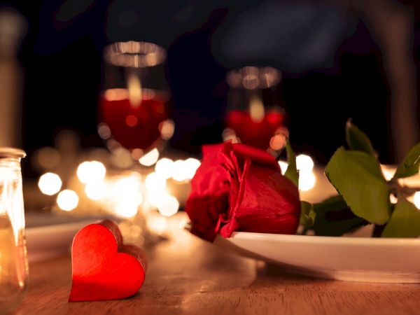 A romantic table setting with a red rose on a plate, heart decor, candles, and glasses of wine on a softly lit dinner table.