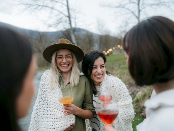Four friends outdoors at a gathering, smiling and holding colorful drinks while chatting with one person partially visible in the foreground.