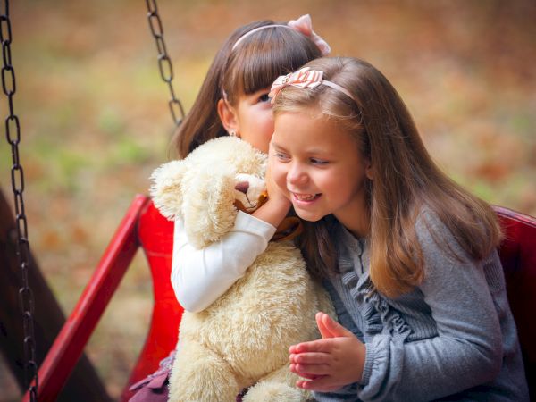 Two little girls sit on a swing, hugging a fluffy teddy bear and smiling warmly in a park.
