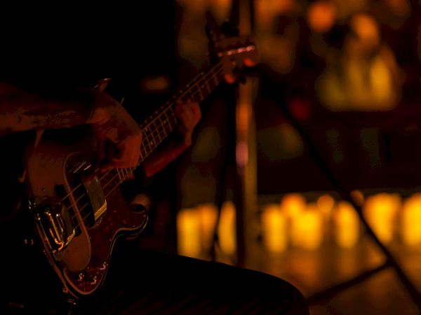 A dimly lit scene with a guitar resting on a chair or stand, blurred warm lights in the background, suggesting a moody concert vibe.