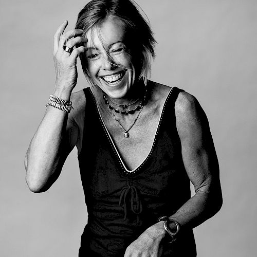 A smiling woman wearing a sleeveless top, layered necklaces and bracelets, mid-laugh, black-and-white portrait.