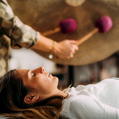 A woman lying down while a person plays a gong with pink mallets.