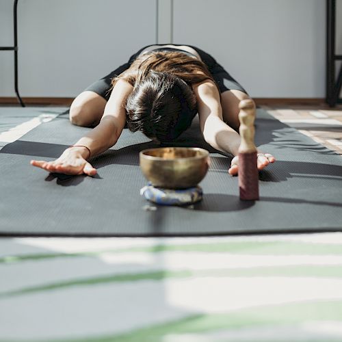 Person in a yoga pose on a mat, arms stretched forward, head down, facing bowls and blocks in a sunlit room, peaceful exercise scene.