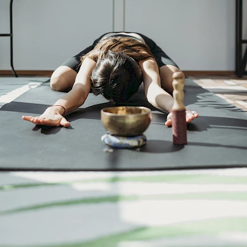 Person in a yoga pose on a mat, arms stretched forward, head down, facing bowls and blocks in a sunlit room, peaceful exercise scene.