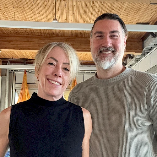 A smiling couple posing for a photo indoors, woman in a sleeveless black top and man in a light gray knit sweater. 140 characters, ending with a period.