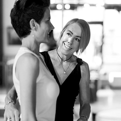 Two women smiling and chatting closely, one with short dark hair and a sleeveless top, the other with a black dress and necklace, in a casual indoor setting.
