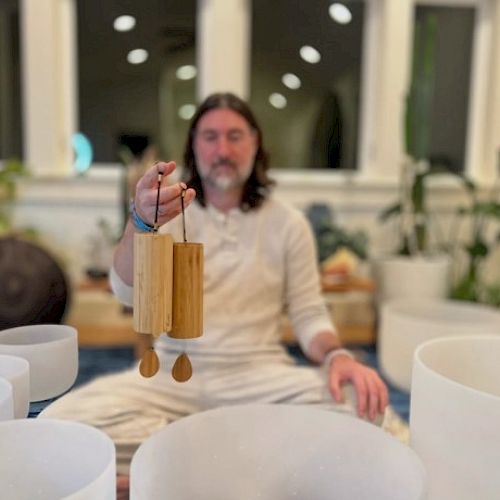 A bearded man in white robes sits cross-legged indoors, holding a wind chime or bamboo chime, surrounded by white bowls or bowls and plants.